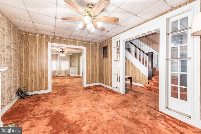 carpeted spare room with a paneled ceiling, a ceiling fan, wood walls, baseboards, and stairs