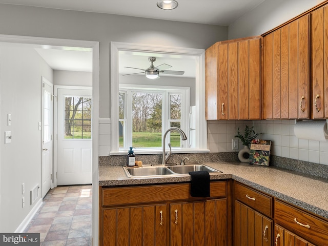 kitchen featuring brown cabinetry, a ceiling fan, a sink, baseboards, and backsplash