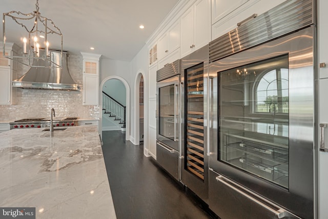 kitchen with light stone counters, arched walkways, tasteful backsplash, white cabinetry, and wall chimney range hood