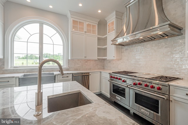 kitchen with light stone counters, appliances with stainless steel finishes, white cabinets, a sink, and wall chimney exhaust hood