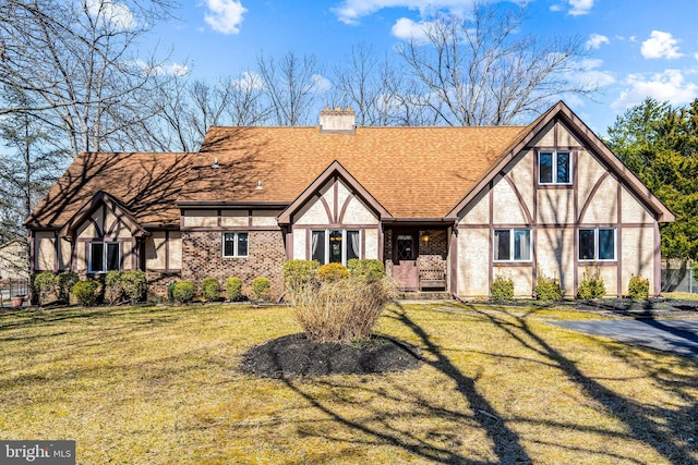 tudor home with brick siding, a front yard, roof with shingles, and a chimney