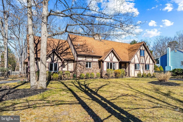 english style home with brick siding, a shingled roof, a front yard, and fence