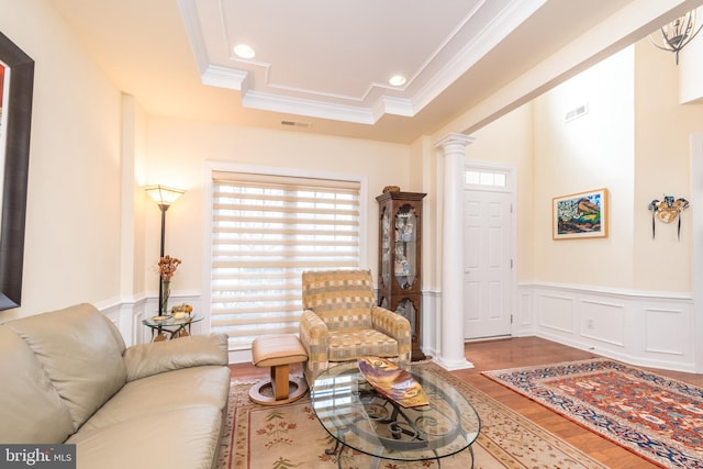 living room featuring wood finished floors, a healthy amount of sunlight, wainscoting, a tray ceiling, and ornate columns