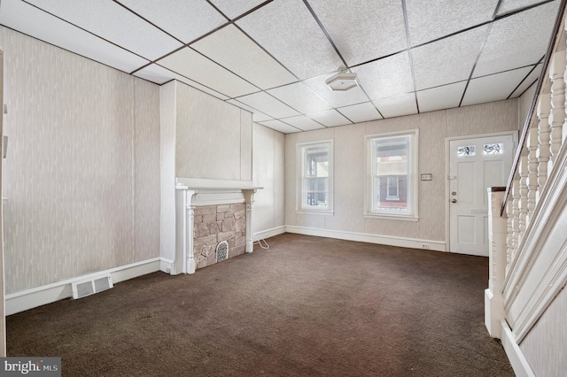 unfurnished living room with stairway, visible vents, a stone fireplace, a paneled ceiling, and dark carpet