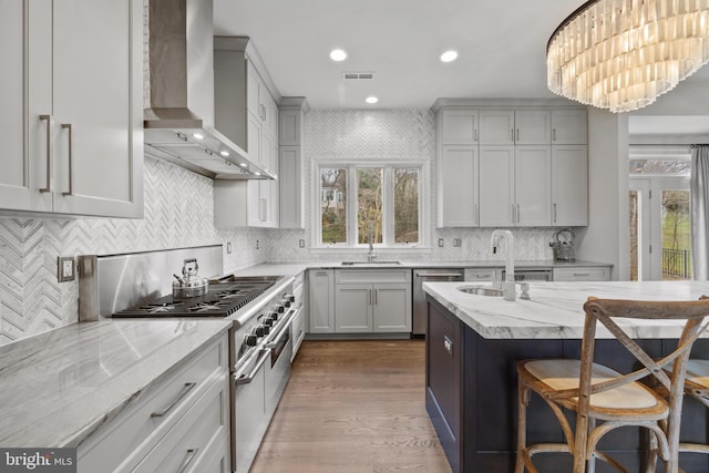 kitchen featuring a kitchen bar, a sink, light stone counters, stainless steel appliances, and wall chimney range hood