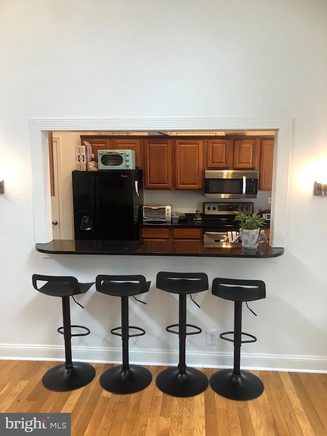 kitchen featuring light wood-type flooring, a kitchen bar, dark countertops, stainless steel appliances, and brown cabinetry