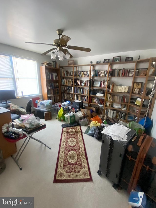 living area featuring a ceiling fan, wall of books, and carpet floors