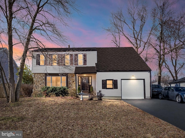 colonial-style house with an attached garage, stone siding, a shingled roof, and aphalt driveway