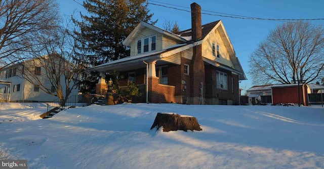 view of snowy exterior featuring a chimney, a storage unit, an outdoor structure, a porch, and brick siding