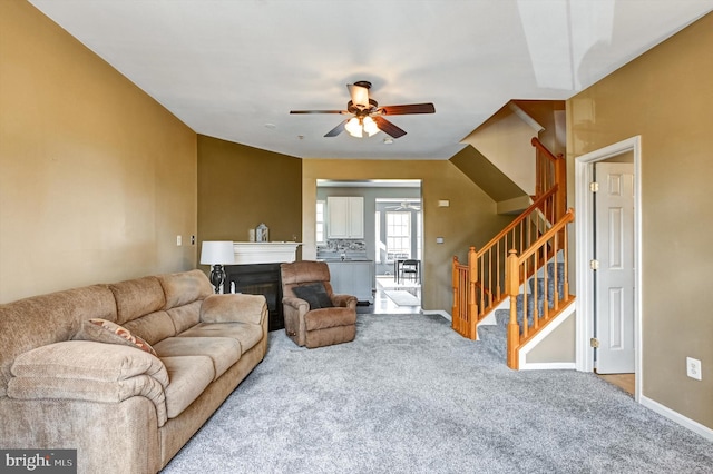 living area featuring carpet flooring, a ceiling fan, baseboards, stairway, and a glass covered fireplace