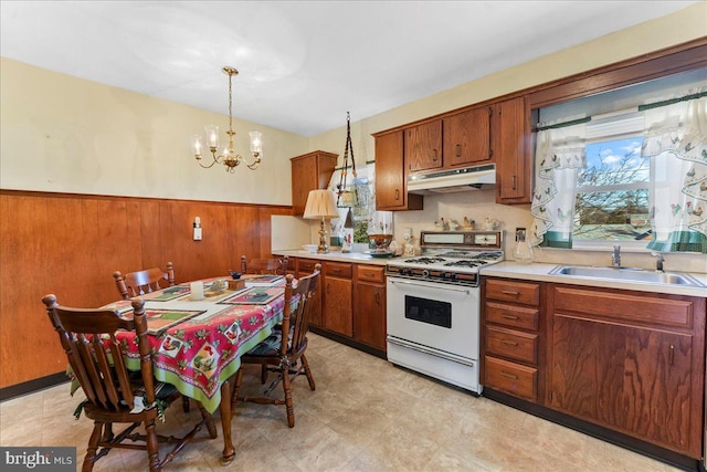 kitchen featuring white gas stove, under cabinet range hood, wainscoting, brown cabinets, and a sink