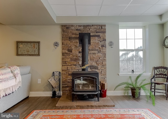 living area with a drop ceiling, a wood stove, baseboards, and wood finished floors