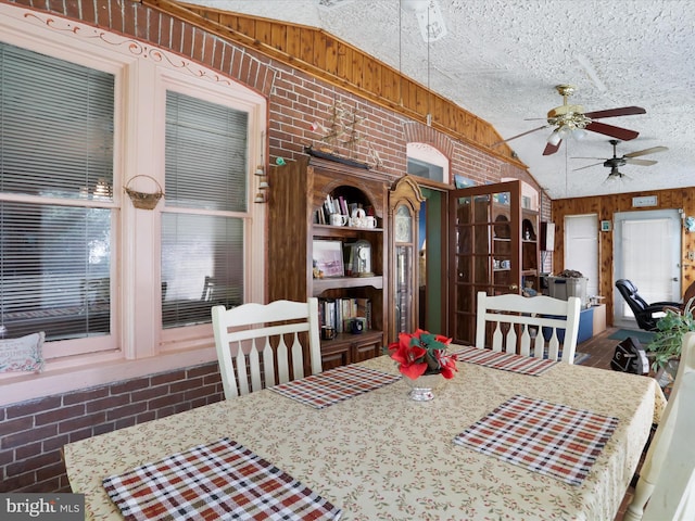 dining area featuring lofted ceiling, a textured ceiling, wooden walls, brick wall, and a ceiling fan