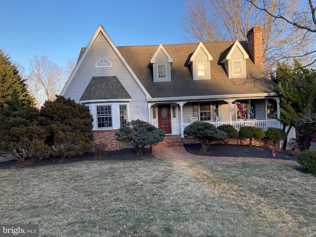 cape cod home featuring brick siding, roof with shingles, a chimney, a porch, and a front yard