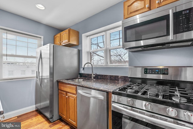 kitchen with a sink, light wood-style floors, appliances with stainless steel finishes, brown cabinetry, and dark stone countertops