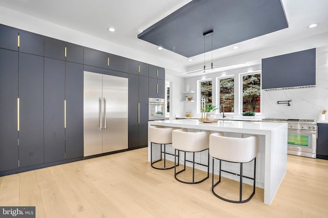 kitchen with light wood finished floors, a raised ceiling, premium appliances, light countertops, and a kitchen bar