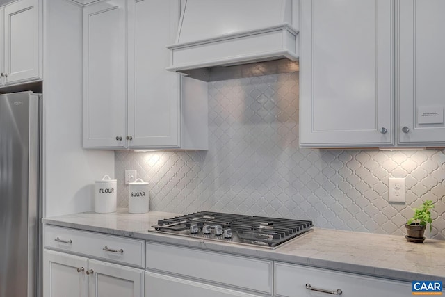 kitchen featuring light stone counters, custom range hood, decorative backsplash, appliances with stainless steel finishes, and white cabinetry