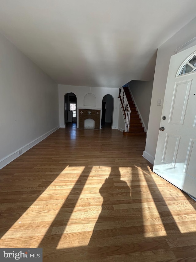 unfurnished living room featuring wood finished floors, baseboards, a fireplace, arched walkways, and stairs