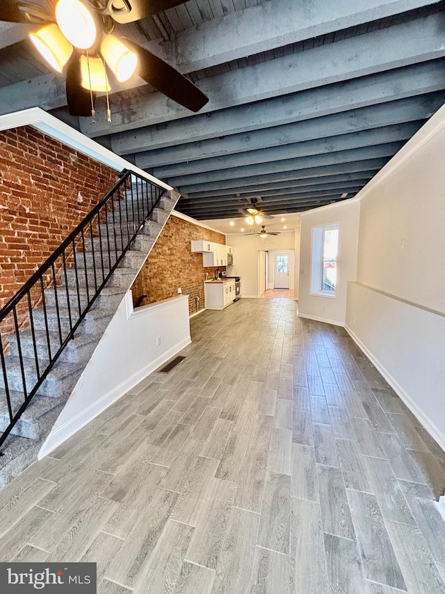 unfurnished living room with ceiling fan, brick wall, wood finished floors, stairs, and beam ceiling