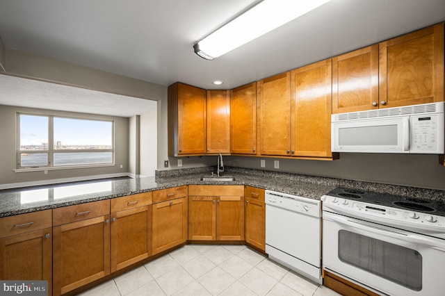 kitchen with white appliances, light tile patterned floors, brown cabinets, dark stone countertops, and a sink