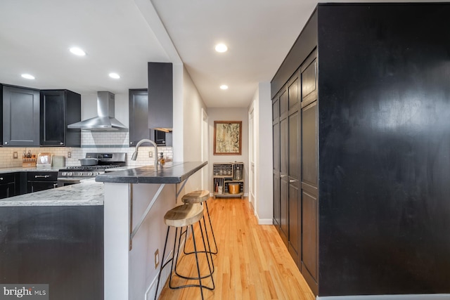 kitchen featuring wall chimney range hood, a kitchen breakfast bar, backsplash, gas stove, and light wood finished floors