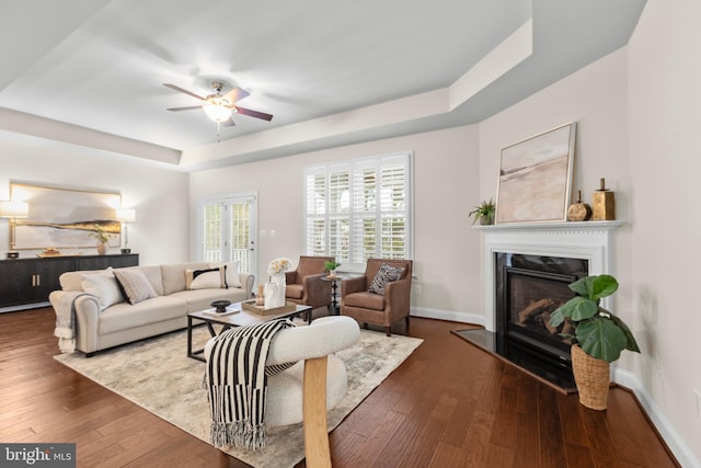 living room with a tray ceiling, dark wood-style flooring, a ceiling fan, a glass covered fireplace, and baseboards