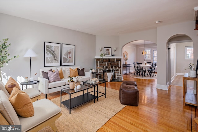 living area featuring arched walkways, a stone fireplace, light wood finished floors, and baseboards