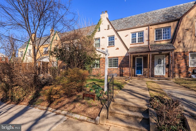 view of front of home with brick siding, a chimney, stucco siding, covered porch, and a high end roof