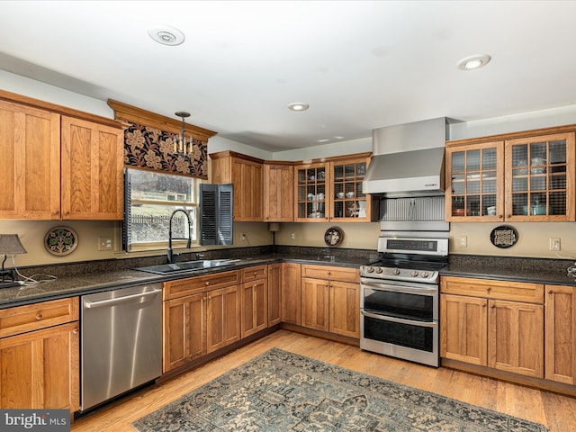 kitchen with light wood finished floors, appliances with stainless steel finishes, brown cabinetry, wall chimney exhaust hood, and a sink