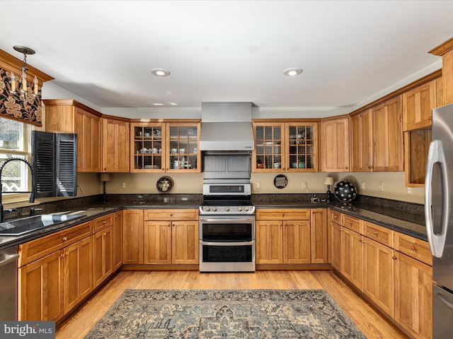 kitchen featuring light wood finished floors, a sink, stainless steel appliances, glass insert cabinets, and wall chimney exhaust hood