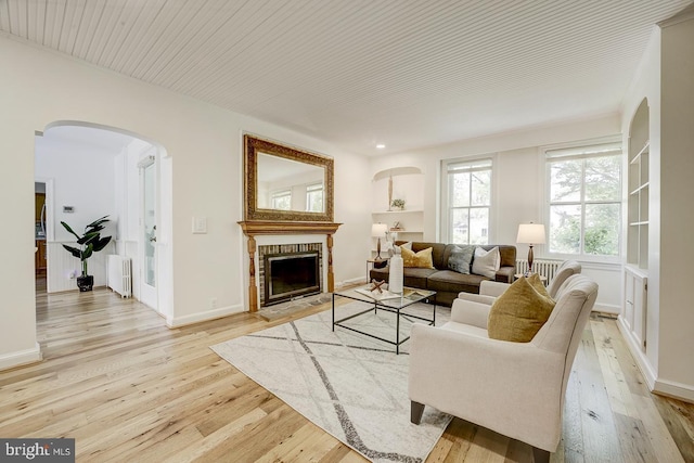 living area with a fireplace, light wood-style floors, baseboards, radiator heating unit, and arched walkways