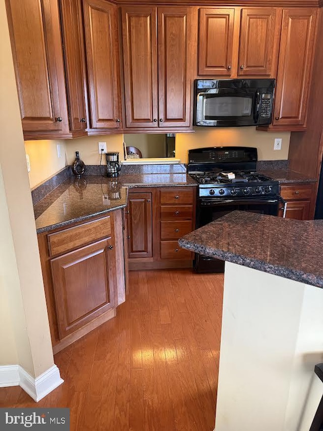 kitchen featuring light wood finished floors, baseboards, dark stone counters, brown cabinets, and black appliances