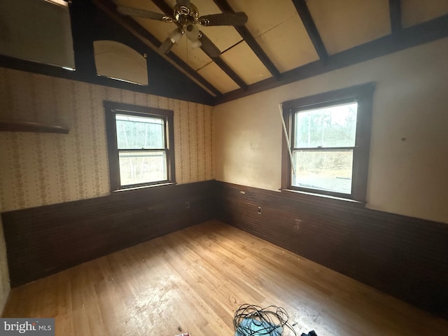 empty room featuring light wood-type flooring, a wainscoted wall, vaulted ceiling with beams, and a ceiling fan