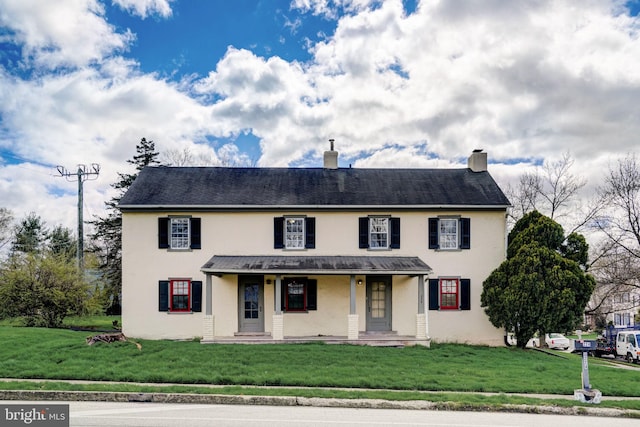 colonial home featuring covered porch, a chimney, a front yard, and stucco siding