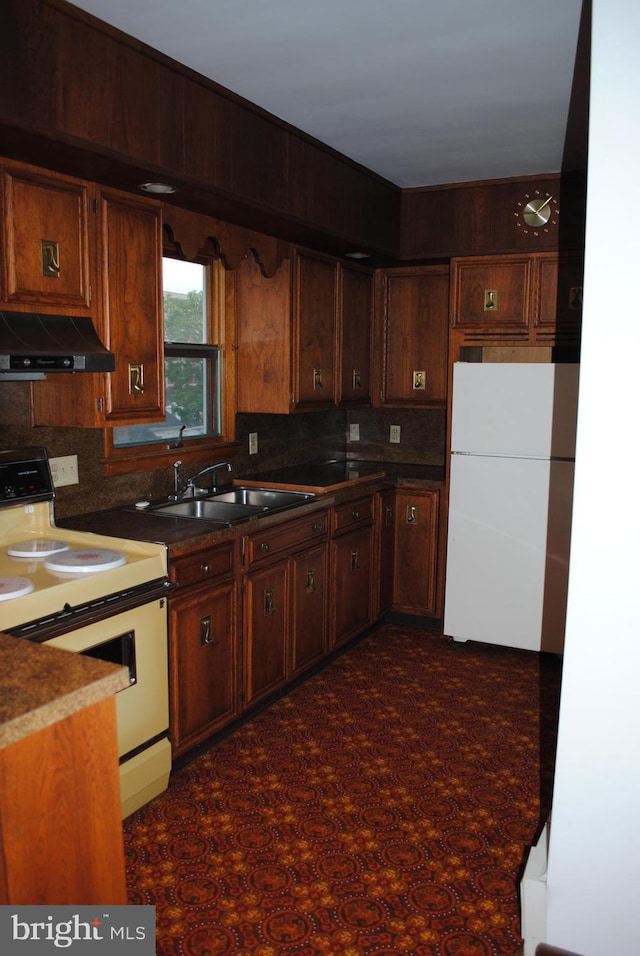 kitchen featuring under cabinet range hood, white appliances, a sink, decorative backsplash, and dark countertops