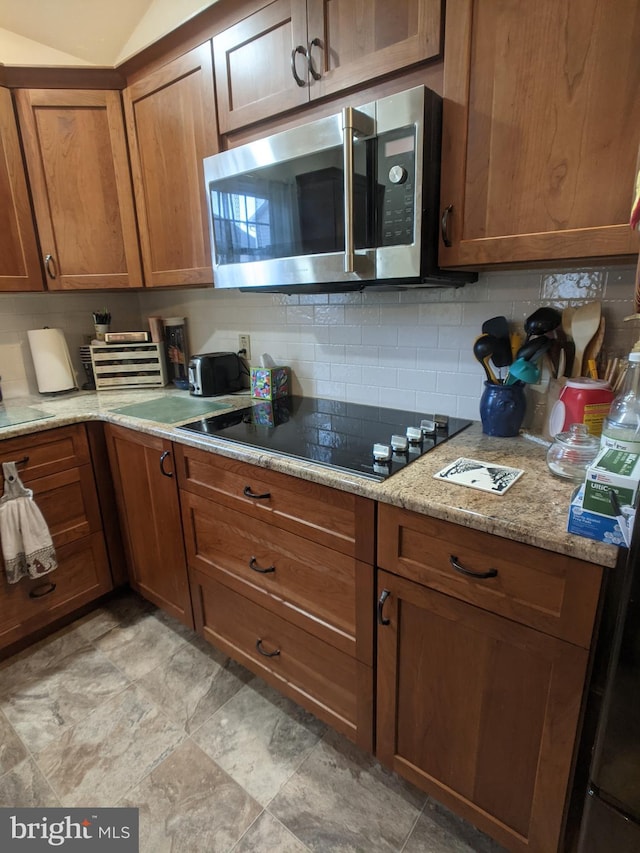 kitchen with light stone counters, black electric cooktop, stainless steel microwave, backsplash, and brown cabinetry