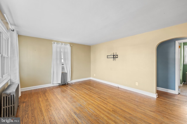 empty room featuring arched walkways, radiator, baseboards, and wood-type flooring