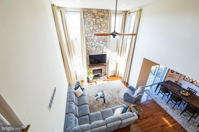 living area featuring ceiling fan, a stone fireplace, wood finished floors, and a towering ceiling