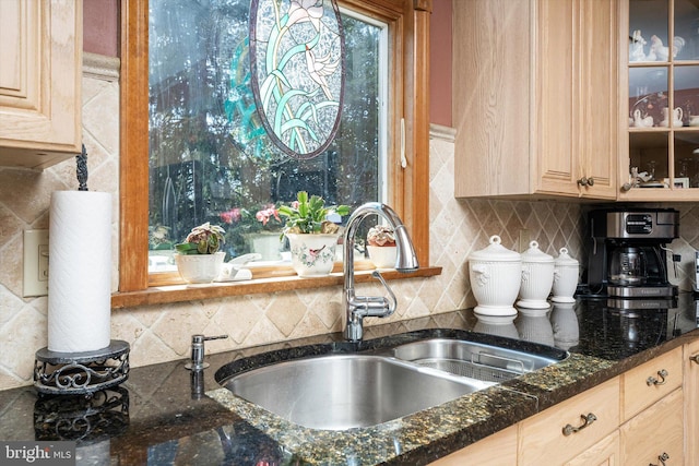 kitchen with light brown cabinetry, a sink, dark stone countertops, backsplash, and glass insert cabinets