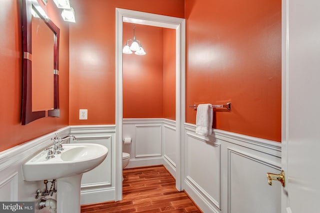 half bath with toilet, wood finished floors, wainscoting, a decorative wall, and a chandelier