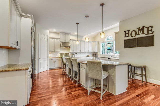 kitchen with wood finished floors, a center island with sink, decorative backsplash, under cabinet range hood, and a kitchen bar