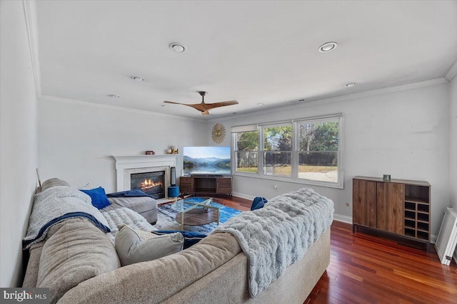 living room featuring baseboards, a glass covered fireplace, ceiling fan, dark wood-type flooring, and crown molding