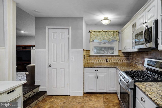 kitchen with a wainscoted wall, stone counters, glass insert cabinets, appliances with stainless steel finishes, and white cabinetry