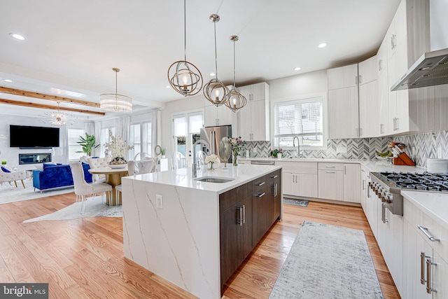 kitchen with light wood-style flooring, a center island with sink, a sink, appliances with stainless steel finishes, and wall chimney exhaust hood