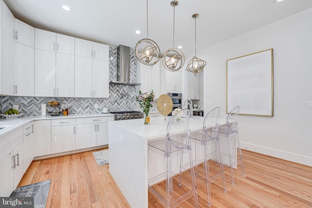 kitchen with wall chimney range hood, pendant lighting, light wood-type flooring, decorative backsplash, and a kitchen breakfast bar