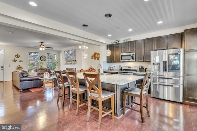 kitchen featuring light stone counters, dark brown cabinetry, stainless steel appliances, a breakfast bar, and dark wood-style flooring