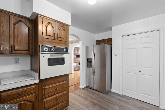 kitchen featuring arched walkways, light wood-style flooring, oven, light countertops, and stainless steel fridge with ice dispenser