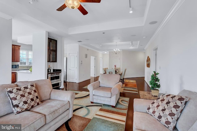 living area featuring a tray ceiling, ornamental molding, wood finished floors, baseboards, and ceiling fan with notable chandelier