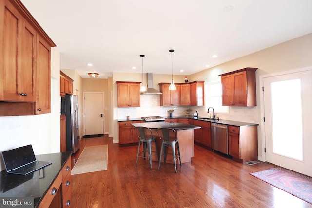 kitchen with a sink, a kitchen island, dark wood-style floors, stainless steel appliances, and wall chimney range hood