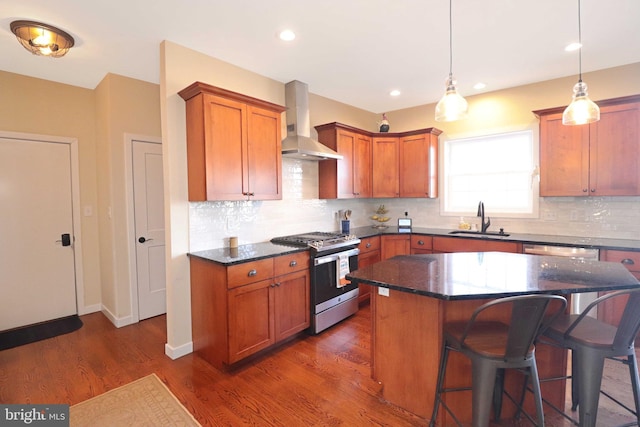 kitchen featuring dark wood finished floors, a sink, appliances with stainless steel finishes, a kitchen bar, and wall chimney range hood